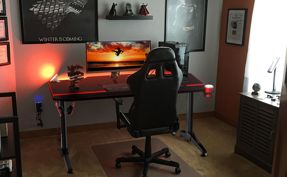 The Vitesse 55-inch gaming desk in front of a wall in a room, with the charging stack, a gaming monitor and a desktop on it. On the wall are mounted two paintings, and mini figures including Superman and Batman. In front of the desk is a racing style gaming chair